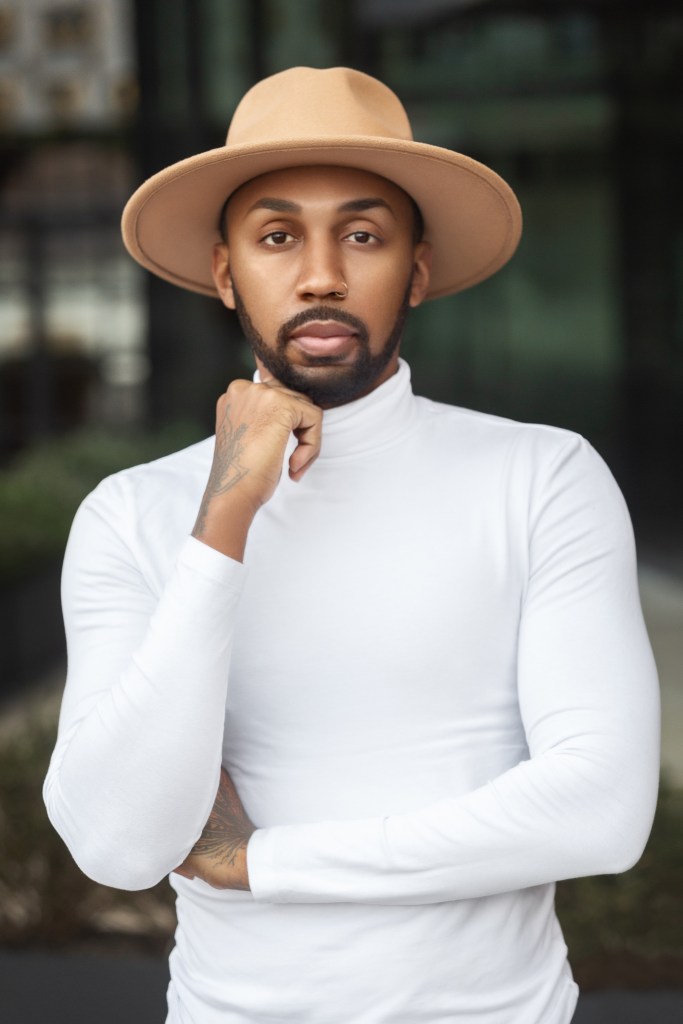 A photograph of the author of "All Boys Aren't Blue", George M. Johnson. They are wearing a khaki hat and a white shirt.