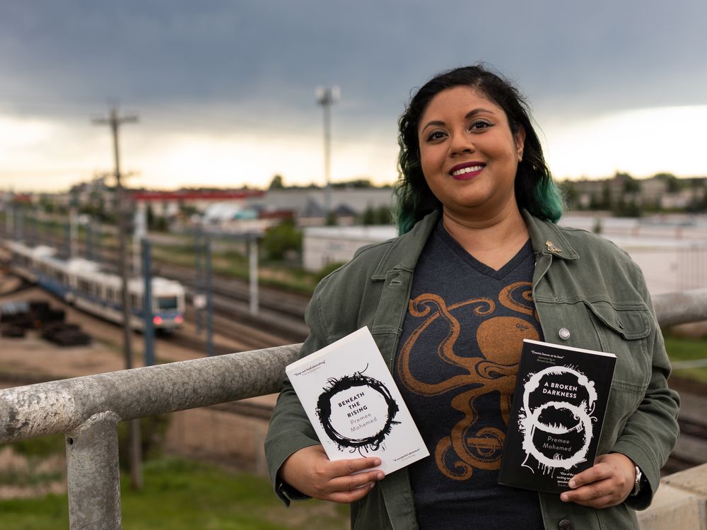 Photograph of the author, Premee Mohamed, holding up a copy of Beneath the Rising and it's sequel, A Broken Darkness